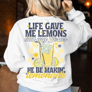 May include: White sweatshirt with a graphic on the back that says "LIFE GAVE ME LEMONS but my Jesus HE BE MAKIN' lemonade." The graphic includes a glass of lemonade, lemons, and stars in yellow and blue.