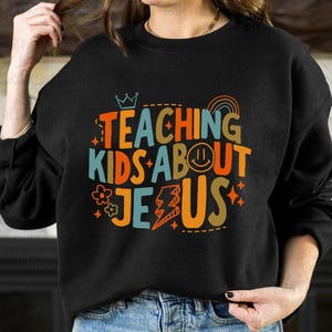 Puede incluir: Sudadera negra con la frase "TEACHING KIDS ABOUT JESUS" en letras coloridas y divertidas. El dise&ntilde;o incluye una corona, un arco&iacute;ris, una carita sonriente, una flor y un rayo. Un estilo casual y c&oacute;modo.