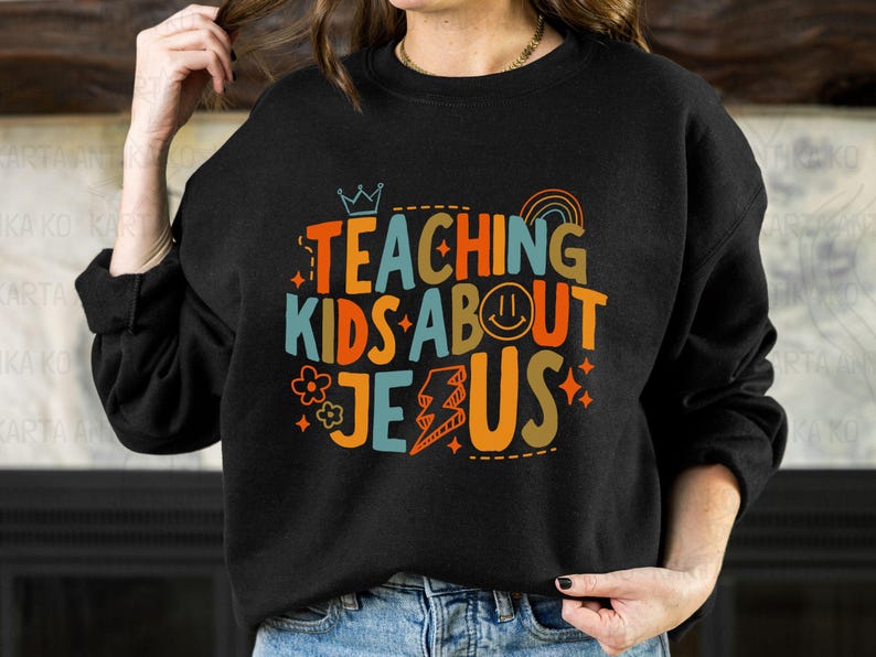 Puede incluir: Sudadera negra con la frase "TEACHING KIDS ABOUT JESUS" en letras coloridas y divertidas. El dise&ntilde;o incluye una corona, un arco&iacute;ris, una carita sonriente, una flor y un rayo. Un estilo casual y c&oacute;modo.