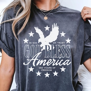 May include: A charcoal grey short-sleeve t-shirt featuring a white graphic print of an eagle surrounded by stars. The text reads: IN GOD WE TRUST, GOD BLESS America, and 250 YEARS OF FREEDOM. The shirt has a crew neckline and a washed-out fabric finish.