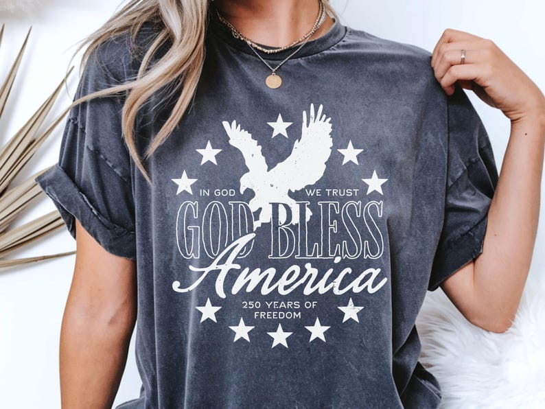 May include: A charcoal grey short-sleeve t-shirt featuring a white graphic print of an eagle surrounded by stars. The text reads: IN GOD WE TRUST, GOD BLESS America, and 250 YEARS OF FREEDOM. The shirt has a crew neckline and a washed-out fabric finish.