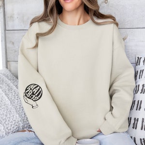 May include: A light beige sweatshirt with a black graphic on the left sleeve. The graphic is a circle with the text "MATT 28:19" inside. The circle is being held by a hand.