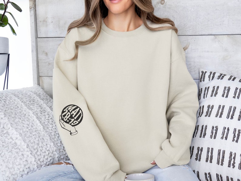 May include: A light beige sweatshirt with a black graphic on the left sleeve. The graphic is a circle with the text "MATT 28:19" inside. The circle is being held by a hand.