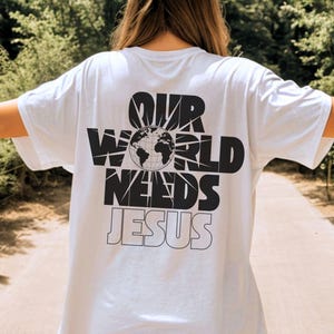 May include: White t-shirt with the text "OUR WORLD NEEDS JESUS" in black, featuring a globe graphic. The shirt is a casual style, suitable for everyday wear.