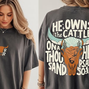 May include: Dark gray t-shirt with a graphic design featuring a bull skull, desert landscape, and the text "He Owns the Cattle on a Thousand Hills Psalm 50:10." A smaller version of the design is on the front.