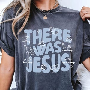 May include: Dark gray t-shirt with the text "THERE WAS JESUS" in light blue, accompanied by butterfly and flower graphics. Additional text includes "in the waiting, in the healing, in the hurting, in the searching."