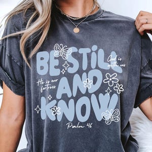 May include: Dark gray t-shirt with the words "BE STILL AND KNOW" in light blue, adorned with butterfly and flower graphics. The text "He is our fortress" and "Psalm 46" are also included.