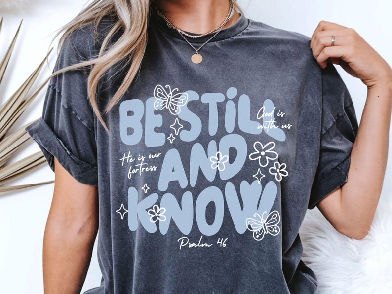 May include: Dark gray t-shirt with the words "BE STILL AND KNOW" in light blue, adorned with butterfly and flower graphics. The text "He is our fortress" and "Psalm 46" are also included.