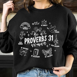 以下が含まれることがあります： 「Be a Proverbs 31 woman」と書かれた白いグラフィックデザインが施された黒いスウェットシャツ。王冠、虹、鳩、蝶、花、聖書などのイラストが描かれています。グラフィックには、「Seek the Lord」、「Be Honest」、「Stay Strong」、「Stay Humble」、「Be Virtuous」、「Keep Serving」、「Be Disciplined」、「Be Faithful」などのフレーズも含まれています。