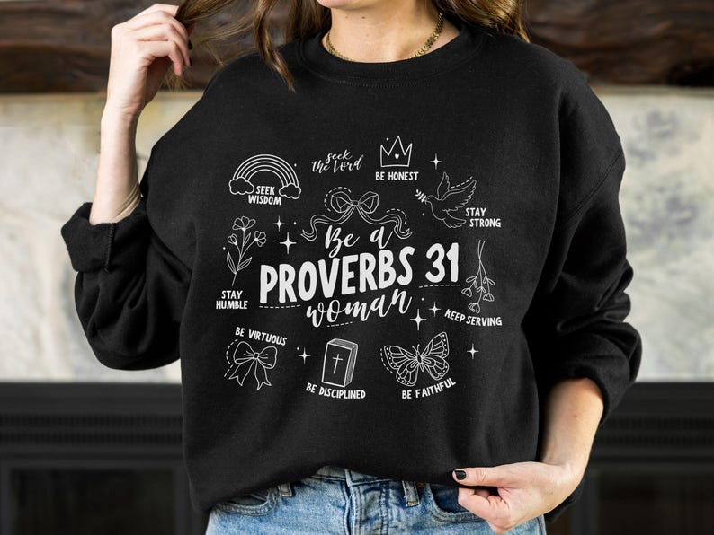 以下が含まれることがあります： 「Be a Proverbs 31 woman」と書かれた白いグラフィックデザインが施された黒いスウェットシャツ。王冠、虹、鳩、蝶、花、聖書などのイラストが描かれています。グラフィックには、「Seek the Lord」、「Be Honest」、「Stay Strong」、「Stay Humble」、「Be Virtuous」、「Keep Serving」、「Be Disciplined」、「Be Faithful」などのフレーズも含まれています。