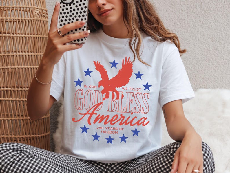 May include: A white t-shirt featuring a red eagle silhouette surrounded by a circle of blue stars. The text reads: IN GOD WE TRUST, GOD BLESS America, 250 YEARS OF FREEDOM. The design uses red and blue ink on a plain white short-sleeved cotton shirt.