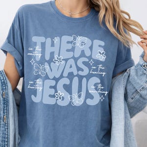 May include: A blue t-shirt with the words "THERE WAS JESUS" in large, light blue letters. The design includes butterflies, flowers, and the phrases "in the waiting," "in the healing," "in the hurting," and "in the searching" in white script.