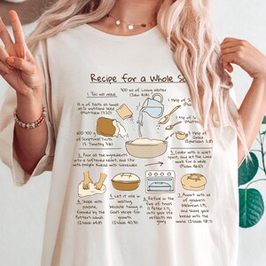 May include: Cream-colored t-shirt featuring a recipe graphic titled "Recipe for a Whole Soul." The graphic includes illustrations of ingredients and steps for baking bread, with Bible verses. The shirt is worn by a person making a peace sign.