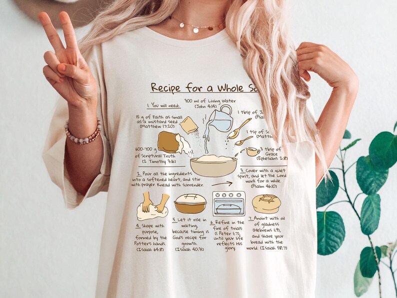May include: Cream-colored t-shirt featuring a recipe graphic titled "Recipe for a Whole Soul." The graphic includes illustrations of ingredients and steps for baking bread, with Bible verses. The shirt is worn by a person making a peace sign.