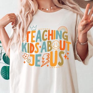Puede incluir: Camiseta color crema con el texto "TEACHING KIDS ABOUT JESUS" en letras coloridas y divertidas. El dise&ntilde;o incluye una corona, un arco&iacute;ris, una carita sonriente, un rayo, flores y estrellas.