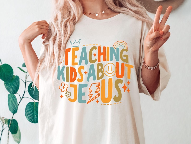 Puede incluir: Camiseta color crema con el texto "TEACHING KIDS ABOUT JESUS" en letras coloridas y divertidas. El dise&ntilde;o incluye una corona, un arco&iacute;ris, una carita sonriente, un rayo, flores y estrellas.
