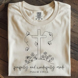 Puede incluir: Camiseta color crema con un diseño de cruz negra, mariposas y flores. El texto "fearfully and wonderfully made" y "Salmo 139:14" está impreso debajo. Fabricado por Comfort Colors.
