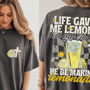 May include: Dark gray t-shirt with a lemon and cross graphic on the front. The back of the shirt features a lemonade graphic with the text "LIFE GAVE ME LEMONS but my Jesus HE BE MAKIN' lemonade."