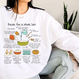 May include: White sweatshirt with a recipe graphic titled "Recipe for a Whole Soul." The graphic includes illustrated steps and ingredients like living water, mustard seed, and joy. The sweatshirt is a comfortable, casual style.
