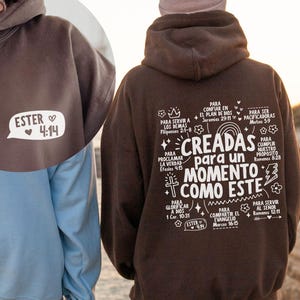 May include: A brown hoodie with the text "ESTER 4:14" in a white speech bubble on the front. The back of the hoodie features a white graphic with Spanish text and illustrations. The hoodie is made of a soft material.