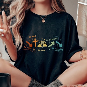 May include: A black t-shirt with a graphic design of a cross, rainbow, and arrows. The text on the shirt reads "He died", "He came", "He rose", "He ascended", and "He's coming back!"