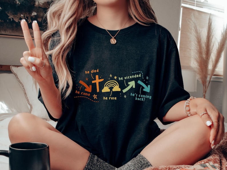 May include: A black t-shirt with a graphic design of a cross, rainbow, and arrows. The text on the shirt reads "He died", "He came", "He rose", "He ascended", and "He's coming back!"