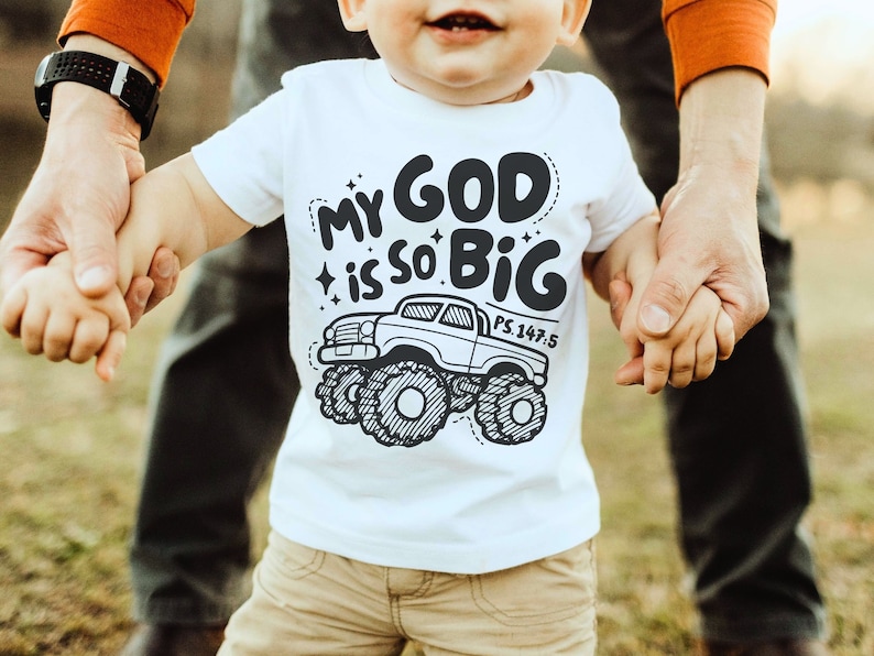 Puede incluir: Una camiseta blanca para ni&ntilde;os peque&ntilde;os con una ilustraci&oacute;n en blanco y negro de un cami&oacute;n monstruo. El texto en la camiseta dice "My God is so Big" con la referencia b&iacute;blica "PS. 147:5".