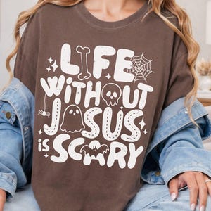 May include: A brown t-shirt featuring white graphic text that reads "LIFE WITHOUT JESUS IS SCARY." The design includes illustrations of a bone, a spider web, a skull, a ghost, a spider, and a bat, all arranged around the lettering on the front.