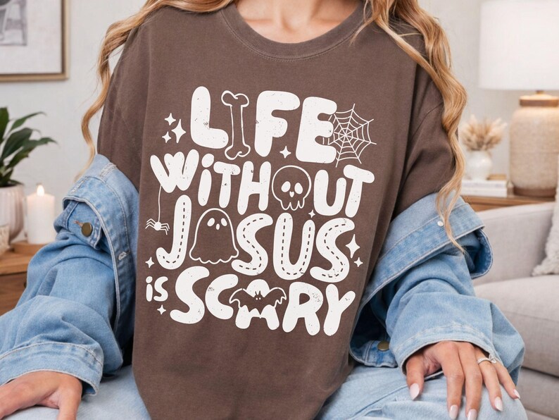 May include: A brown t-shirt featuring white graphic text that reads "LIFE WITHOUT JESUS IS SCARY." The design includes illustrations of a bone, a spider web, a skull, a ghost, a spider, and a bat, all arranged around the lettering on the front.