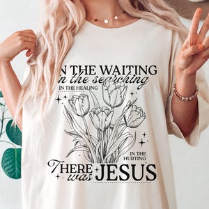 May include: Cream-colored t-shirt featuring a black and white floral design with the words "IN THE WAITING, IN THE SEARCHING, IN THE HEALING, IN THE HURTING, THERE WAS JESUS."