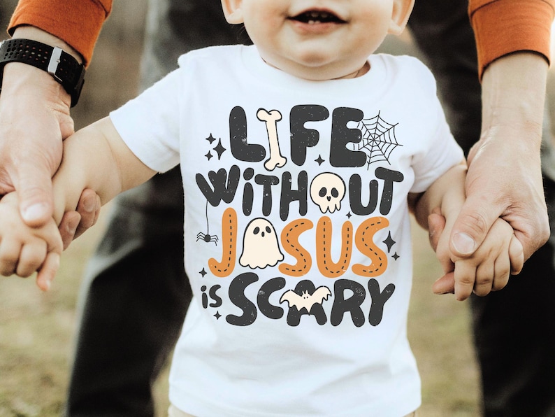May include: A white short-sleeved t-shirt featuring the black and orange text "LIFE WITHOUT JESUS IS SCARY" with graphic illustrations of a bone, a spider web, a skull, a ghost, a spider, and a bat integrated into the lettering design.