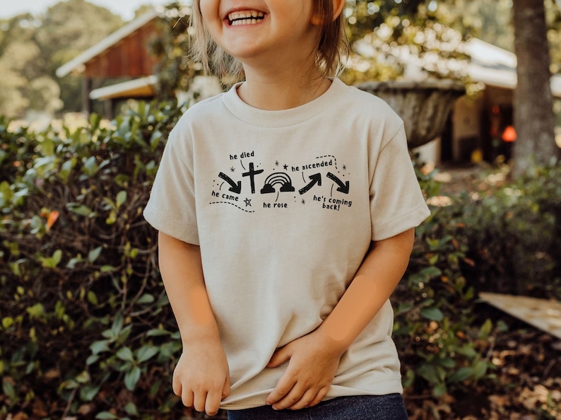May include: A light beige toddler t-shirt with a black graphic design featuring a cross, stars, a rainbow, and arrows. The text on the shirt reads "he died", "he came", "he rose", "he ascended", and "he's coming back!"