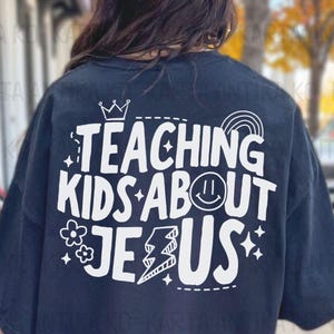 Puede incluir: Camiseta azul marino con un dise&ntilde;o blanco que dice "TEACHING KIDS ABOUT JESUS". El dise&ntilde;o incluye una corona, un arco&iacute;ris, una carita sonriente, flores, un rayo y estrellas. Una prenda informal.