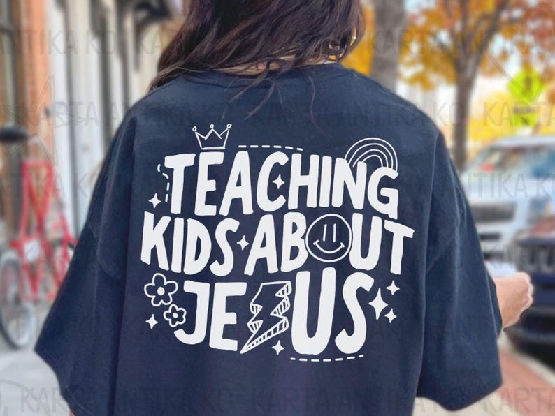 Puede incluir: Camiseta azul marino con un dise&ntilde;o blanco que dice "TEACHING KIDS ABOUT JESUS". El dise&ntilde;o incluye una corona, un arco&iacute;ris, una carita sonriente, flores, un rayo y estrellas. Una prenda informal.