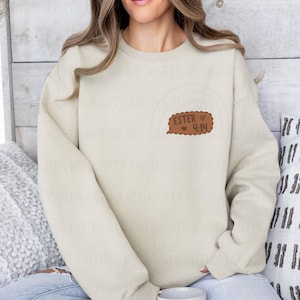 May include: Cream-colored sweatshirt with a brown speech bubble graphic. The speech bubble contains the text "ESTER 4:14" in a handwritten style, with a small heart symbol. The sweatshirt is a casual, long-sleeved garment.