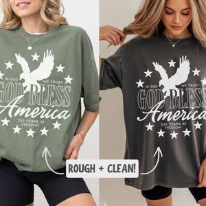 May include: Two oversized t-shirts in olive green and charcoal grey, each featuring a white graphic of an eagle surrounded by stars and the text: IN GOD WE TRUST, GOD BLESS AMERICA, 250 YEARS OF FREEDOM. The shirts have a crew neck and short sleeves.