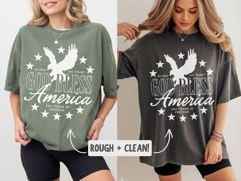 May include: Two oversized t-shirts in olive green and charcoal grey, each featuring a white graphic of an eagle surrounded by stars and the text: IN GOD WE TRUST, GOD BLESS AMERICA, 250 YEARS OF FREEDOM. The shirts have a crew neck and short sleeves.