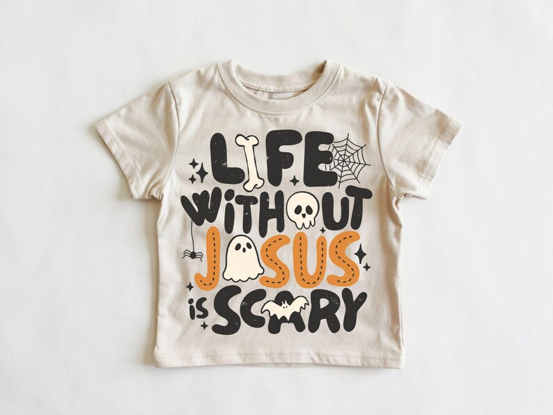 May include: A beige short-sleeve t-shirt featuring black and orange graphic text that reads "LIFE WITHOUT JESUS IS SCARY." The design includes illustrations of a bone, a spider web, a skull, a ghost, a small spider, and a bat.