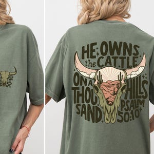 May include: Olive green t-shirt with a bull skull graphic. The front features a small bull skull design with the text "PSALM 50:10". The back displays a larger bull skull with the text "HE OWNS THE CATTLE ON A THOUSAND HILLS PSALM 50:10".