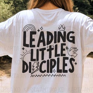 Könnte beinhalten: Weißes T-Shirt mit schwarzem Text, der "LEADING LITTLE DISCIPLES" lautet. Der Text ist von verschiedenen schwarzen und weißen grafischen Elementen umgeben, darunter ein Regenbogen, Sterne, eine Krone, eine Sonne, ein Blitz und eine Taube.