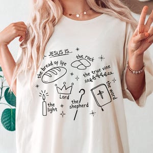 May include: White t-shirt with black hand-drawn illustrations of religious symbols and the text "JESUS IS... the bread of life, the rock, the true vine, the light, Lord, the shepherd, the word".