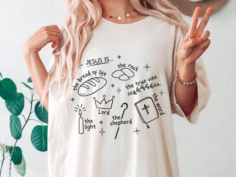 May include: White t-shirt with black hand-drawn illustrations of religious symbols and the text "JESUS IS... the bread of life, the rock, the true vine, the light, Lord, the shepherd, the word".