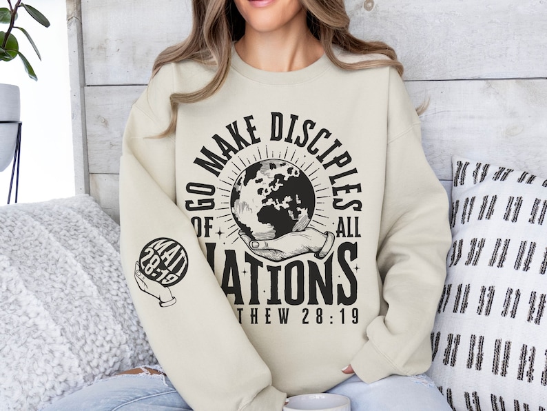 May include: A beige sweatshirt with a black graphic print that says "Go Make Disciples of All Nations" with a globe and a hand holding it. The text "Matthew 28:19" is printed below the graphic. The left sleeve has a smaller graphic of a hand holding a globe with the text "Matt 28:19".