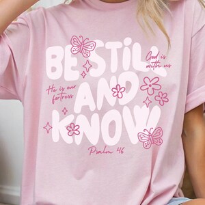May include: A light pink t-shirt with the words "BE STILL AND KNOW" in large white letters. The shirt features pink butterfly and flower graphics, along with the phrases "God is with us" and "He is our fortress". The text "Psalm 46" is also printed on the shirt.