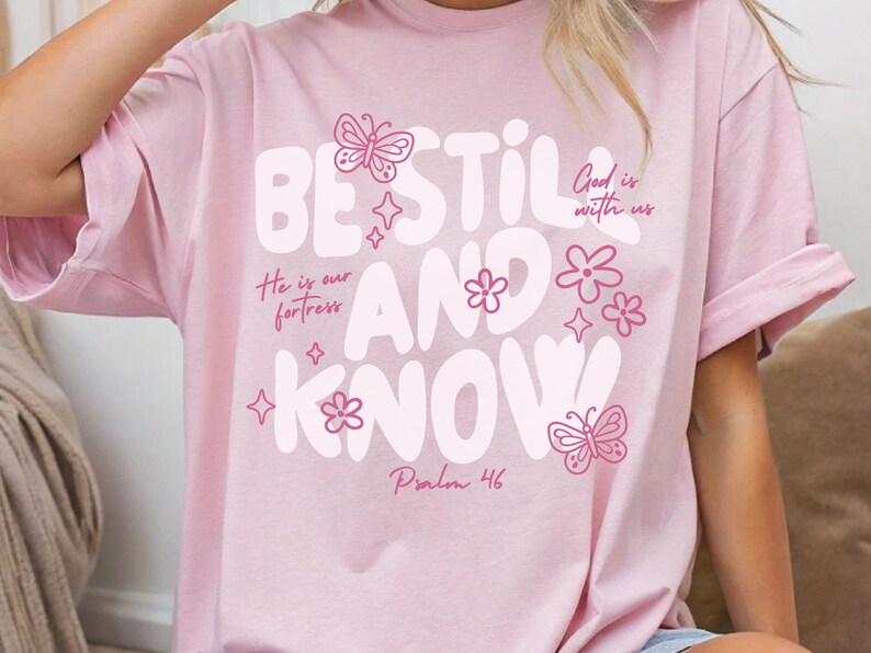 May include: A light pink t-shirt with the words "BE STILL AND KNOW" in large white letters. The shirt features pink butterfly and flower graphics, along with the phrases "God is with us" and "He is our fortress". The text "Psalm 46" is also printed on the shirt.