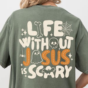 May include: An olive green t-shirt featuring a graphic on the back with the text "LIFE WITHOUT JESUS IS SCARY" in a retro font. The design includes white and orange illustrations of a bone, spider web, skull, ghost, spider, and bat.