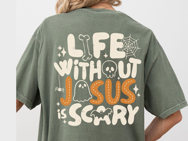 May include: An olive green t-shirt featuring a graphic on the back with the text "LIFE WITHOUT JESUS IS SCARY" in a retro font. The design includes white and orange illustrations of a bone, spider web, skull, ghost, spider, and bat.