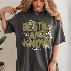 May include: Dark gray t-shirt with the words "BE STILL AND KNOW" in olive green, along with floral and butterfly graphics. The text "He is our fortress" and "God is with us" are also included. Psalm 46 is written at the bottom.