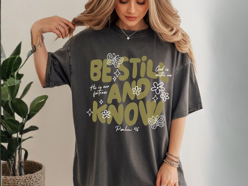 May include: Dark gray t-shirt with the words "BE STILL AND KNOW" in olive green, along with floral and butterfly graphics. The text "He is our fortress" and "God is with us" are also included. Psalm 46 is written at the bottom.