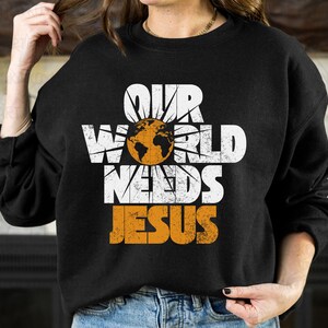 May include: Black sweatshirt with the text "OUR WORLD NEEDS JESUS" in white and orange. The design includes a globe graphic. The sweatshirt is a casual style, suitable for everyday wear.
